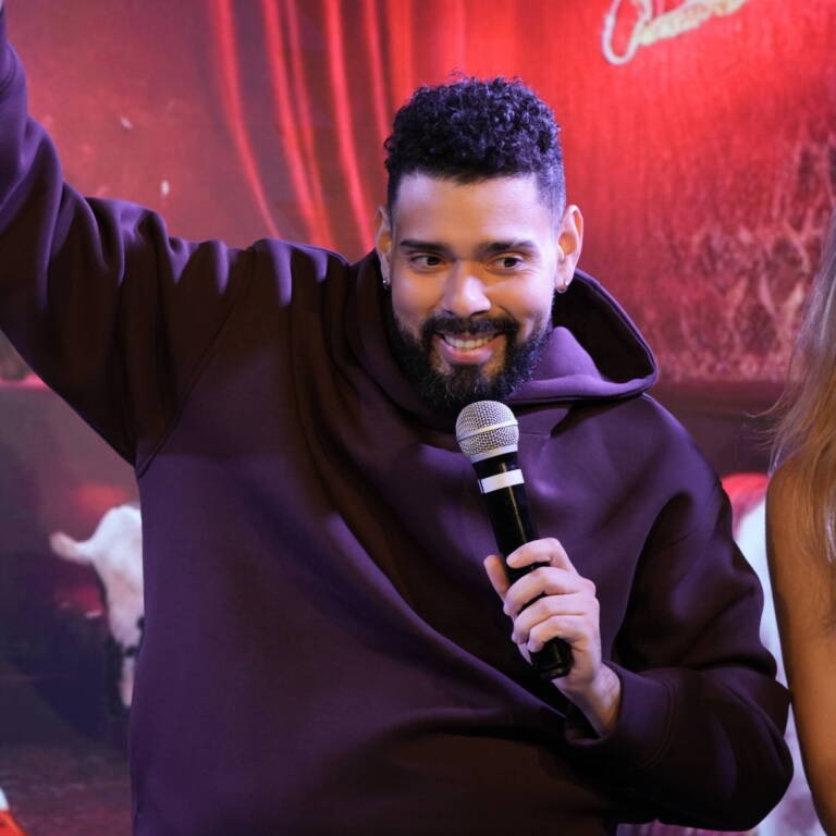 Comedian in a burgundy hoodie raising a hand as he speaks into the microphone