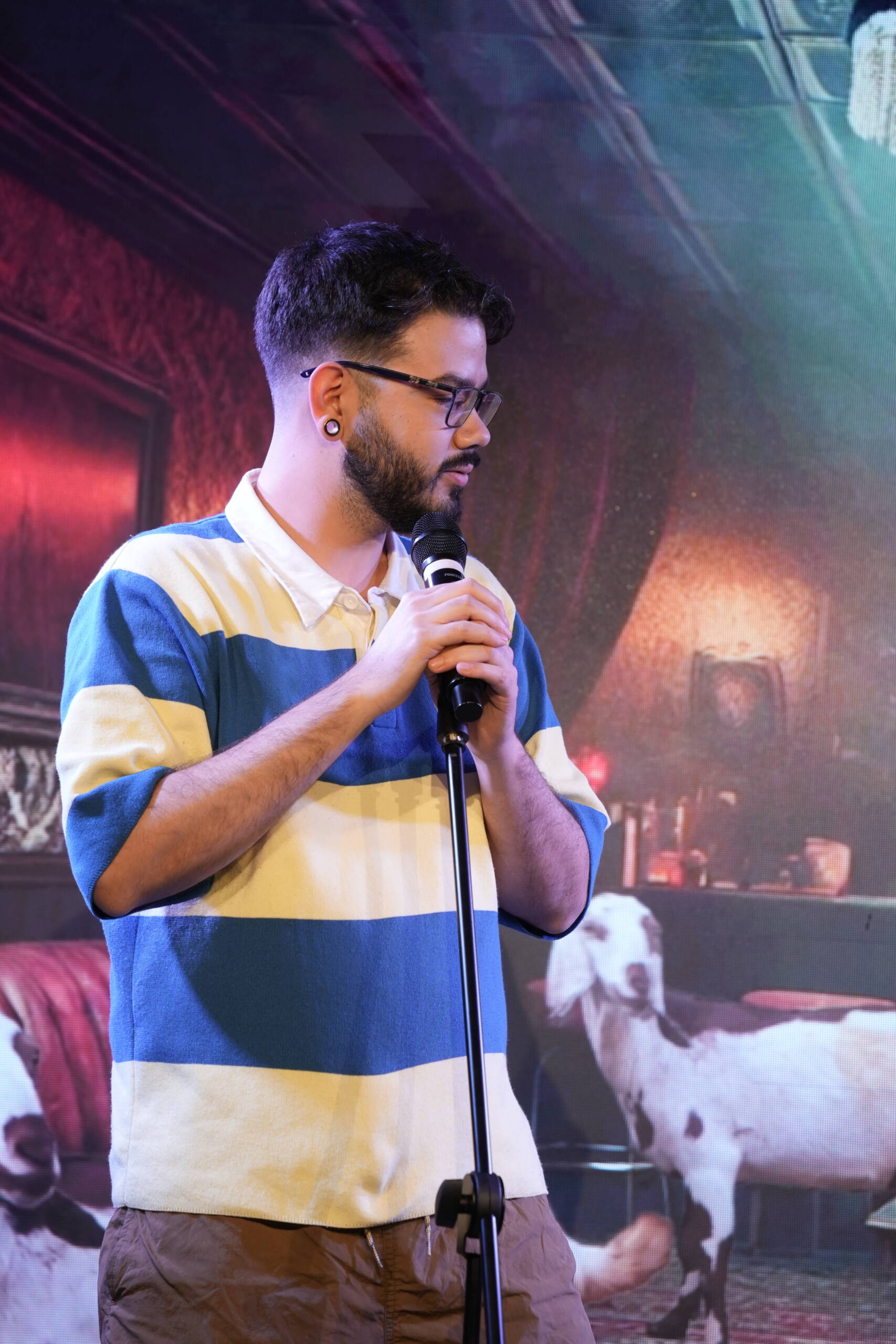 Comedian in a striped polo shirt at the mic stand in front of a red backdrop
