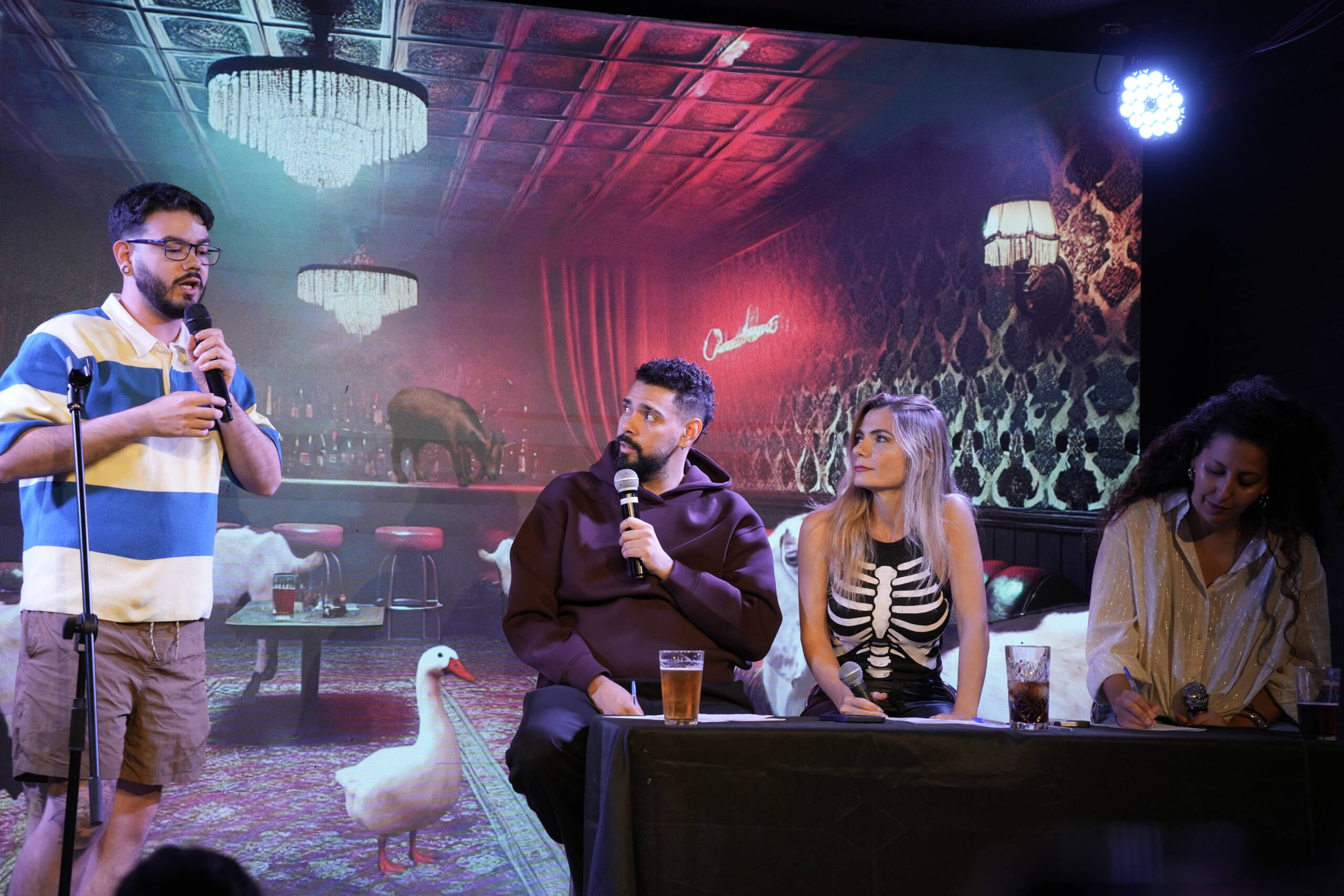 Wide view of a stage dressed with red curtains and chandeliers: a comedian at the mic and three guests seated at a table