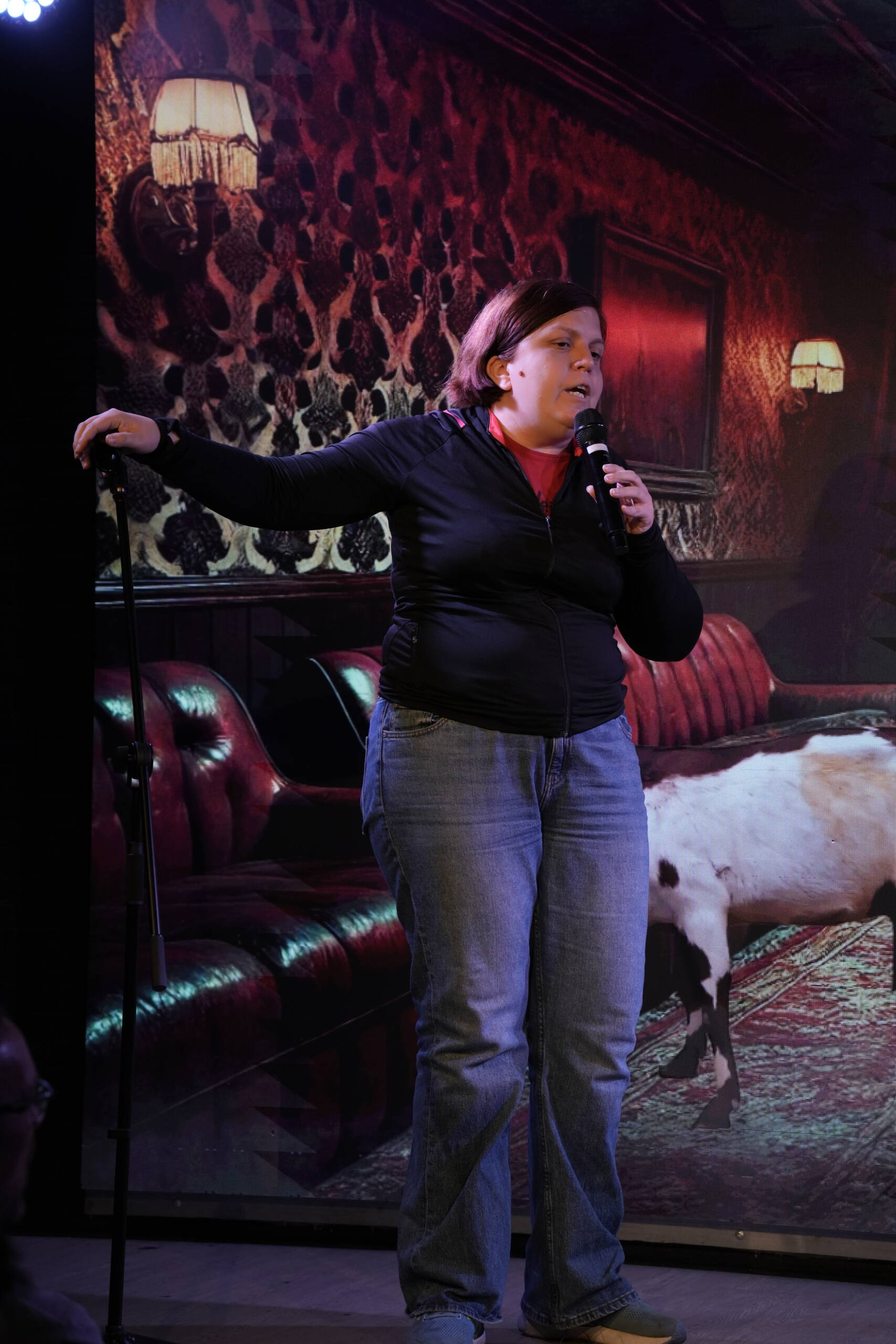 Comedian in a dark jacket gesturing on stage in front of red velvet sofas