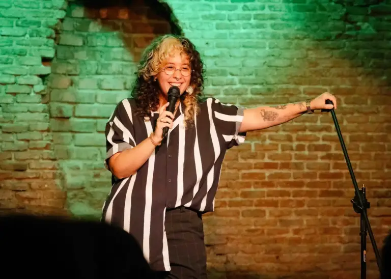 Comedian in a striped shirt performing against a brick wall lit in green