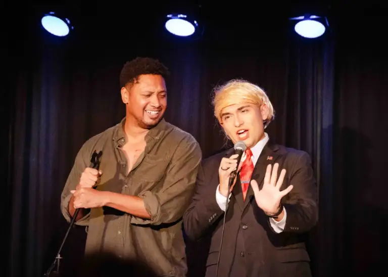 A comedian in a green shirt and a performer in a suit and blond wig sharing the stage