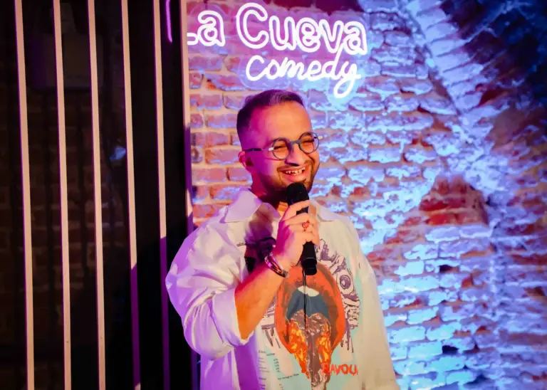 Comedian in glasses holding the microphone against the brick wall at La Cueva Comedy