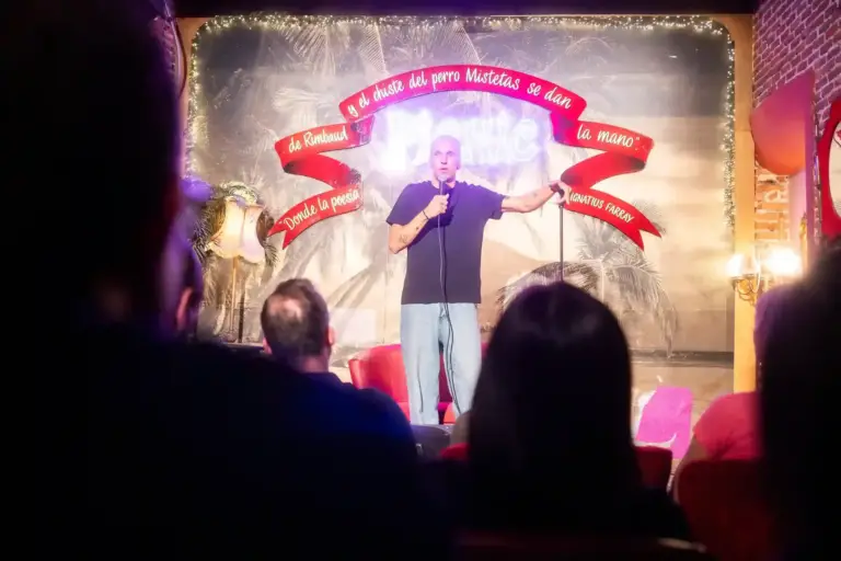 Comedian in a white T-shirt performing with open arms in front of the Picnic backdrop