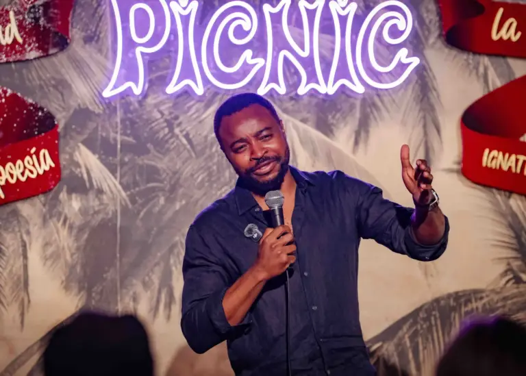 Comedian in a dark shirt gesturing mid-joke under the neon Picnic sign