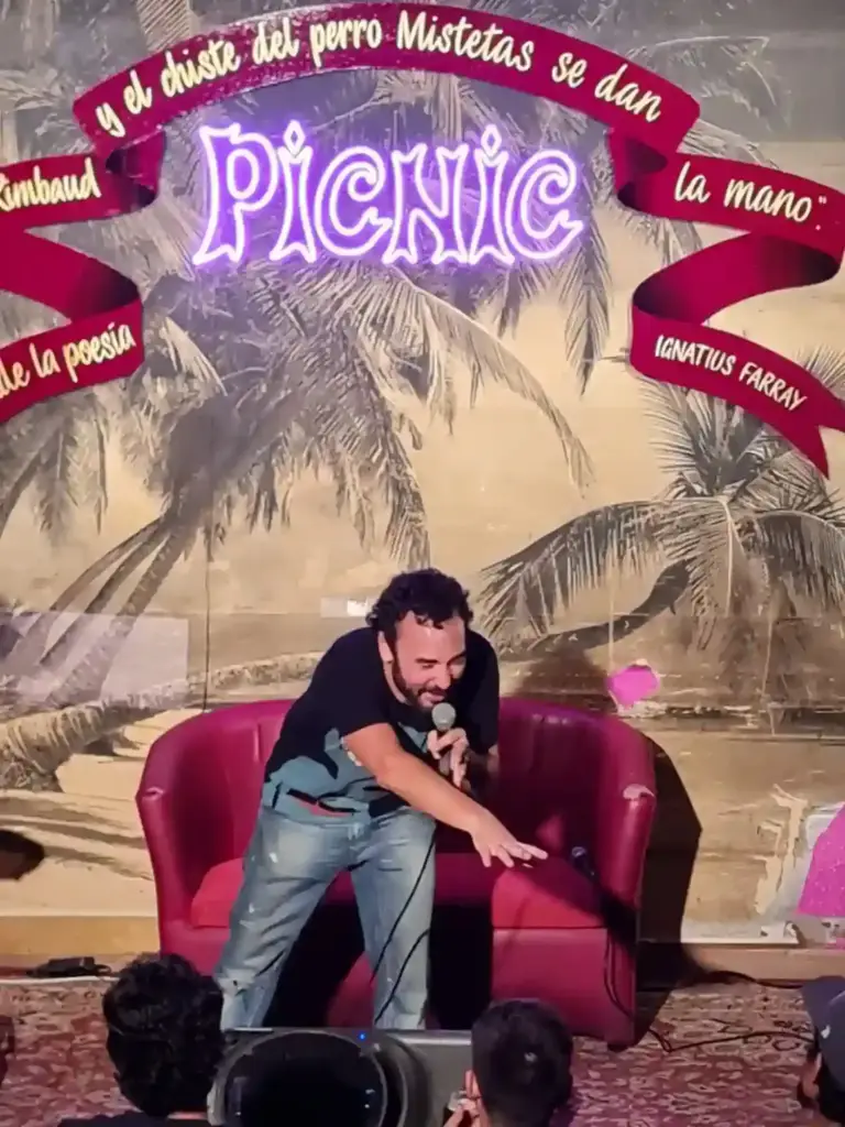 Comedian leaning towards the audience in front of the red armchair on the Picnic stage