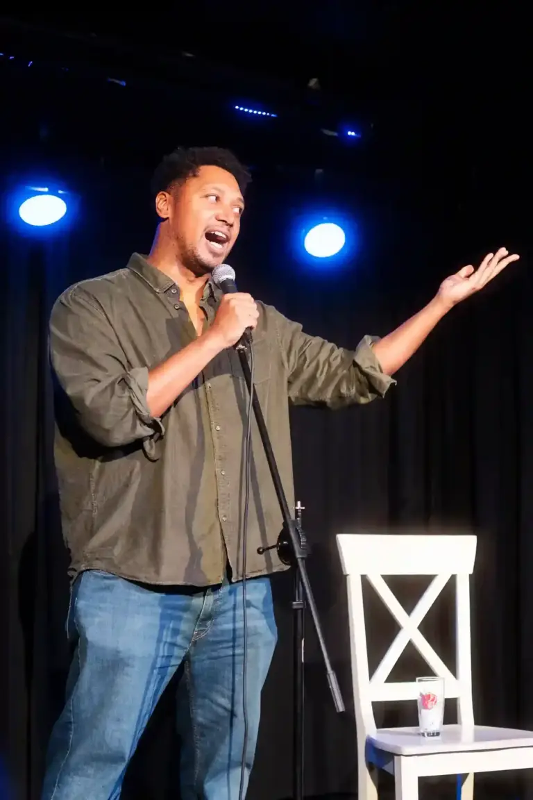Comedian in an olive shirt performing on a dark stage with blue lights