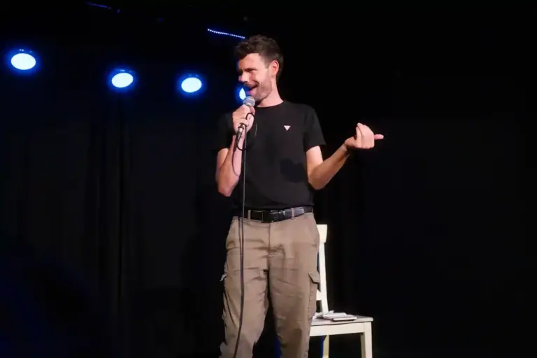 Comedian in a black T-shirt gesturing to the side while performing