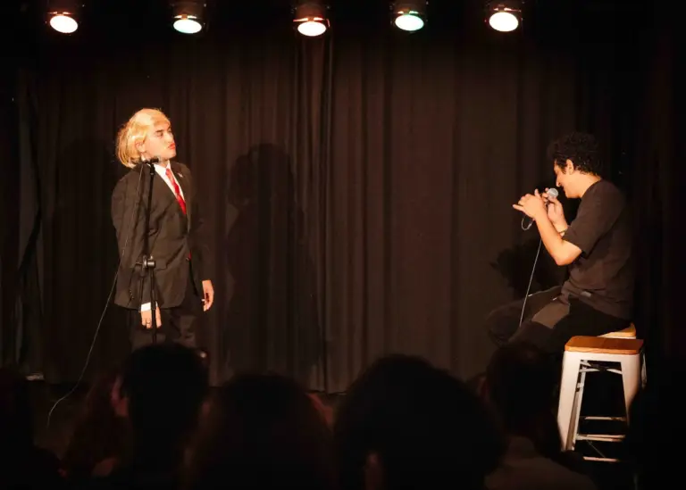 Performer in a suit and blond wig on stage, with a second comedian seated on a stool