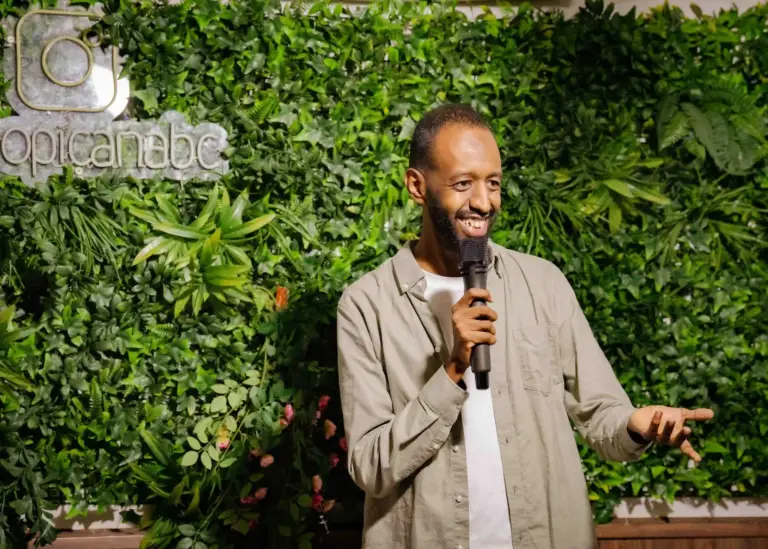 Comedian in a beige overshirt performing in front of the plant wall at Tropicana